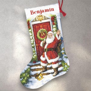 May include: A Christmas stocking with a festive design. It depicts Santa Claus in a red suit, waving in front of a red door adorned with a wreath. The name "Benjamin" is printed at the top, with snow and greenery.
