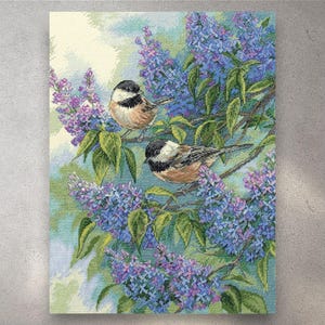 May include: Cross-stitch picture of two small birds on lilac branches. The birds have black and white heads, tan bodies, and grey wings. The lilac flowers are purple and blue, with green leaves. The background is a soft blend of blue and green. A decorative piece.