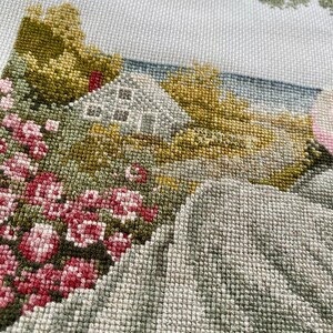 May include: A detailed cross-stitch landscape featuring a white house, pink flowers, and green and yellow trees. The needlework depicts a serene countryside scene with a light blue sky and a textured, hand-stitched appearance.