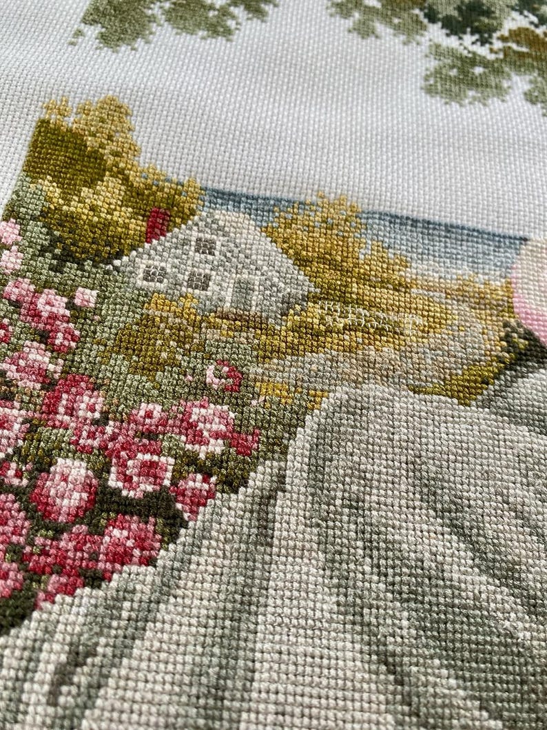 May include: A detailed cross-stitch landscape featuring a white house, pink flowers, and green and yellow trees. The needlework depicts a serene countryside scene with a light blue sky and a textured, hand-stitched appearance.