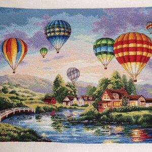 Puede incluir: Paisaje de punto de cruz con globos aerost&aacute;ticos de colores sobre un lago y un pueblo. Los globos presentan patrones vibrantes en rojo, naranja, azul y verde. Un puente blanco cruza el agua.