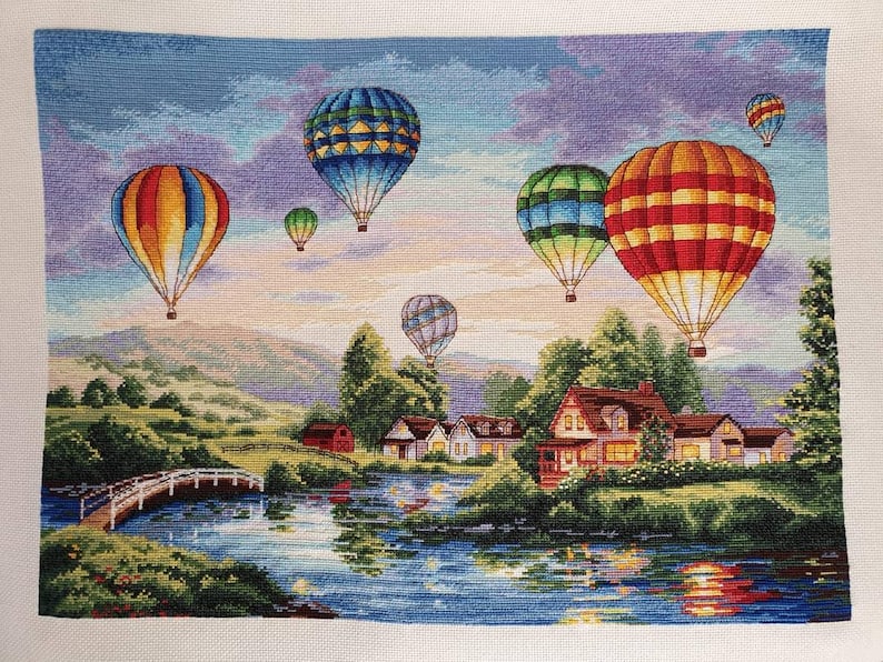 Puede incluir: Paisaje de punto de cruz con globos aerost&aacute;ticos de colores sobre un lago y un pueblo. Los globos presentan patrones vibrantes en rojo, naranja, azul y verde. Un puente blanco cruza el agua.