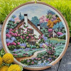 May include: A cross-stitch embroidery of a cottage scene, displayed in a wooden hoop. The cottage features a white exterior, brown roof, and is surrounded by vibrant flowers and greenery. A path leads to the cottage, with a small pond in the foreground.
