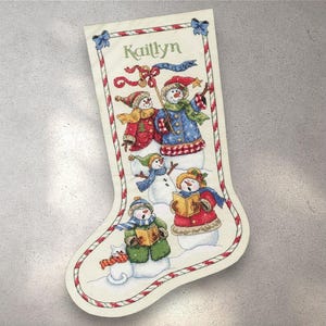 Puede incluir: Un calcetín navideño con un fondo crema, con un diseño festivo de muñecos de nieve cantando villancicos. El nombre "Kaitlyn" está bordado en la parte superior. El calcetín está adornado con un patrón de bastón de caramelo rojo y blanco.