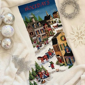 May include: A Christmas stocking with a winter village scene. The stocking features a blue sky with the word "HOLIDAYS" embroidered in red. The village scene includes houses, people, trees, and a horse-drawn sleigh.