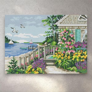 Puede incluir: Obra de arte de punto de cruz que representa una escena costera. Una casa con una contraventana azul claro está rodeada de flores coloridas. El océano tiene veleros y un muelle. Dos gaviotas vuelan en el cielo.