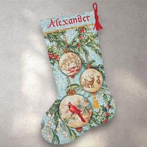 May include: A light blue Christmas stocking with the name "Alexander" embroidered in red. The stocking features three round ornaments with winter scenes and a red cardinal. A red tassel hangs from the top.