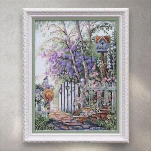 Op de afbeelding: Ingelijst kruissteekkunstwerk dat een serene tuinscène uitbeeldt. De afbeelding toont een witte hek, een bakstenen pad en een vogelhuisje. De tuin is gevuld met paarse en roze bloemen, met een strohoed die aan de poort hangt.