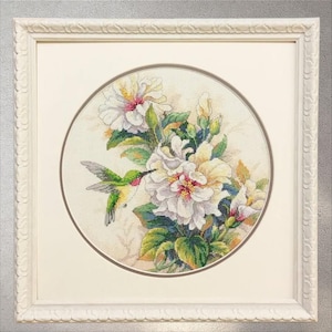 May include: A framed cross-stitch of a hummingbird and white hibiscus flowers. The hummingbird is green and red, near the flowers. The flowers have yellow and pink centres and green leaves. The artwork is in a white frame.