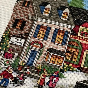 May include: Cross-stitch depiction of a snowy village scene with buildings, people, and a "TOY SHOPPE" sign. The buildings have brick and stone facades, blue shutters, and lit windows. People are bundled up in winter clothing, some pulling a sled.