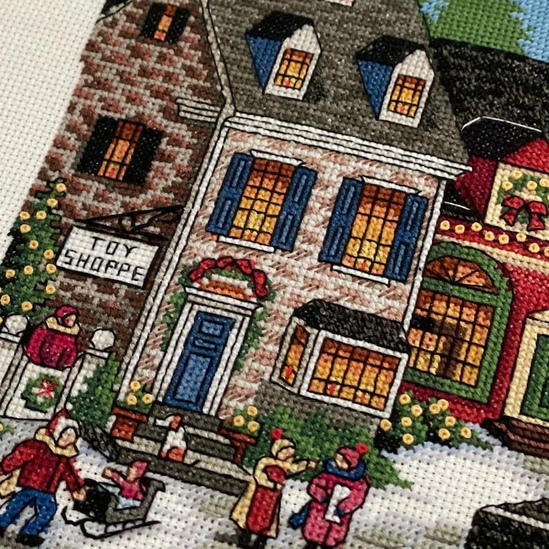 May include: Cross-stitch depiction of a snowy village scene with buildings, people, and a "TOY SHOPPE" sign. The buildings have brick and stone facades, blue shutters, and lit windows. People are bundled up in winter clothing, some pulling a sled.