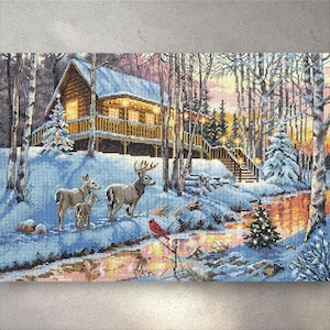 Könnte beinhalten: Kreuzstich einer Winterhütten-Szene mit Hirschen. Die Holzhütte hat Lichter im Inneren. Die Szene zeigt eine Hirschfamilie, einen roten Vogel, Schnee, Bäume und einen Bach. Die Farbpalette ist hauptsächlich blau, weiß, braun und orange.