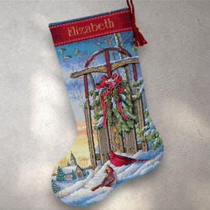 Puede incluir: Un calcetín navideño con una escena invernal que presenta un trineo rojo, cardenales y una iglesia. La banda superior es roja con el nombre "Elizabeth". El calcetín tiene una borla roja y está decorado con una corona y nieve.