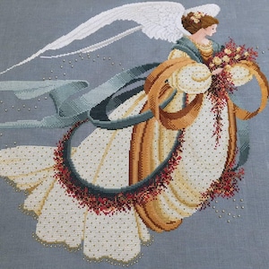 Puede incluir: Imagen de punto de cruz de un ángel con alas blancas, vestido con un vestido crema con detalles dorados y una guirnalda de flores rojas. El ángel sostiene un ramo y está rodeado de cintas azules y marrones. El fondo es gris suave.