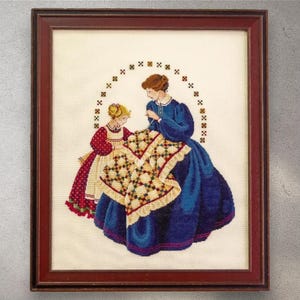 May include: Cross-stitch artwork in a dark wood frame. The image depicts a woman and a young girl sewing a quilt. The woman wears a blue dress, and the girl wears a red dress. The quilt has a yellow and green pattern.