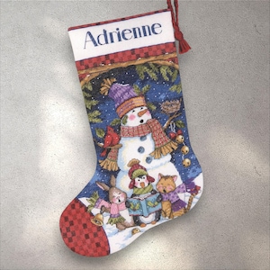 Puede incluir: Un calcetín navideño con una escena de muñeco de nieve y animales. El calcetín presenta un muñeco de nieve con un sombrero morado y una bufanda roja y marrón. El nombre "Adrienne" está impreso en la parte superior. La parte inferior del calcetín tiene un patrón de cuadros rojos y negros.