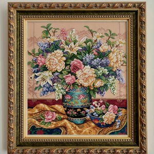 May include: Framed cross-stitch artwork featuring a floral still life. The arrangement includes peonies, roses, lilies, and other flowers in a decorative vase and bowl. The frame is gold with ornate detailing.