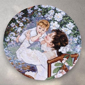 May include: A round needlepoint featuring a mother holding a baby. The mother wears a white dress and sits in a wooden chair. The baby is in a white outfit. The background is a garden with white flowers.