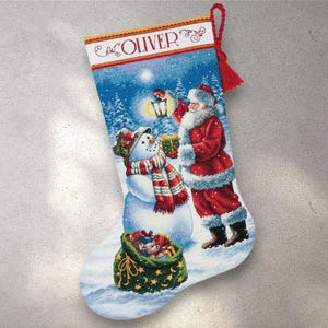 May include: Christmas stocking featuring Santa Claus and a snowman in a snowy scene. The stocking has a red tassel and the name "OLIVER" at the top. Santa is holding a lantern and a gift, while the snowman wears a hat and scarf.