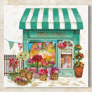 May include: A cross-stitch image of a flower shop with a green and white striped awning. The shop front is teal with the word "BLOOMS" above the window. Baskets and pots of colourful flowers are displayed outside the shop.