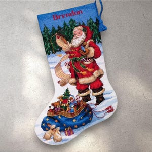 May include: Embroidered Christmas stocking featuring Santa Claus holding a list, with a blue sack filled with toys. The stocking has a blue top with the name "Brendan" and a tassel.