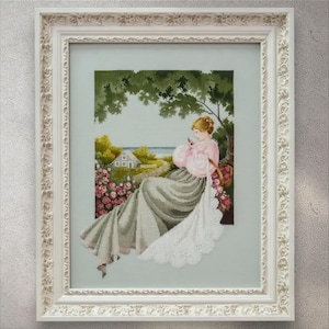 May include: Cross-stitch artwork in a white ornate frame. The artwork depicts a woman in a pink dress seated under a tree, with a house and garden in the background. The scene is framed by a border of pink roses.