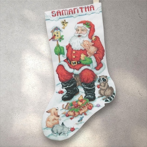 May include: White Christmas stocking with a cross-stitch design featuring Santa Claus, animals, and the name "SAMANTHA" in red letters. Santa is in a red suit, holding a pig and a bird. A raccoon and other animals are also present.