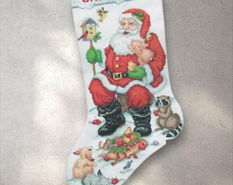 May include: White Christmas stocking with a cross-stitch design featuring Santa Claus, animals, and the name "SAMANTHA" in red letters. Santa is in a red suit, holding a pig and a bird. A raccoon and other animals are also present.