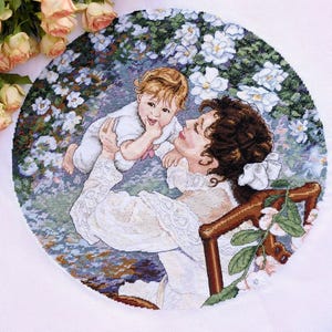 May include: A cross-stitch embroidery depicts a mother holding a baby, surrounded by white flowers. The mother wears a white dress, and the baby is in a white outfit. The artwork is circular, with a floral background and a wooden chair.