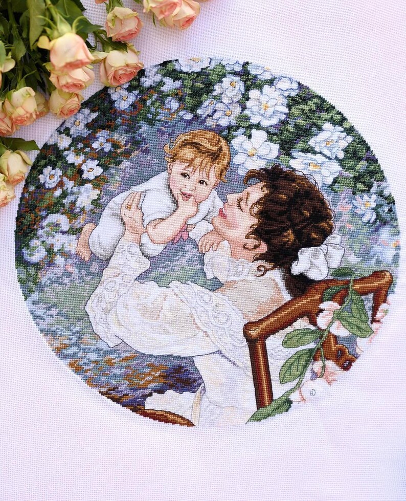 May include: A cross-stitch embroidery depicts a mother holding a baby, surrounded by white flowers. The mother wears a white dress, and the baby is in a white outfit. The artwork is circular, with a floral background and a wooden chair.