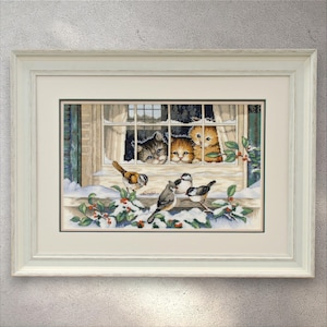 Puede incluir: Obra de arte enmarcada que presenta una escena invernal con tres gatitos mirando por una ventana a pájaros posados en una repisa cubierta de nieve. La obra tiene un marco blanco y representa un ambiente invernal acogedor con bayas de acebo.