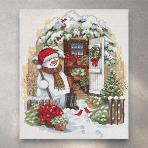 Puede incluir: Una escena invernal en punto de cruz con un muñeco de nieve con gorro y bufanda rojos, sosteniendo una pala. La escena incluye un jardín nevado con una valla de madera, un pequeño árbol de Navidad y poinsettias rojas. Una corona festiva adorna la puerta.