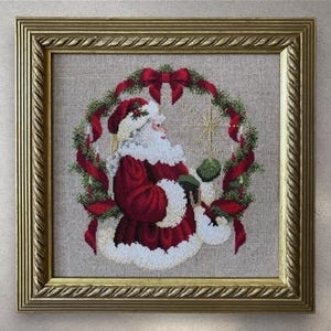 Può includere: Opera d'arte a punto croce incorniciata raffigurante Babbo Natale in abito rosso con finiture bianche, che tiene in mano un oggetto verde. È circondato da una ghirlanda di verde e nastri rossi. La cornice è dorata.