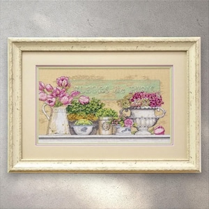 May include: Cross-stitch artwork depicting a floral still life. A white jug holds pink tulips, and a sign reads "Fleurs de Paris." The artwork is framed in a cream-coloured frame.