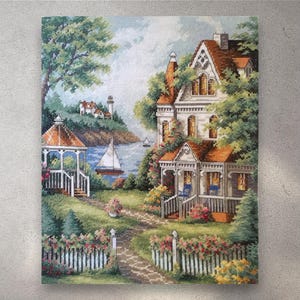 May include: A cross-stitch landscape showing a coastal scene with a large house, gazebo, and sailboat. The house has a red roof and is surrounded by greenery and flowers. A white picket fence lines a path leading to the house.
