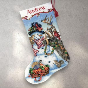 Könnte beinhalten: Weihnachtsstrumpf mit Schneemann, Rentieren und Vögeln. Der Strumpf trägt oben den Namen "Andrew" in roten Buchstaben. Unten befindet sich ein Korb mit Äpfeln und Geschenken. Der Hintergrund ist ein verschneites Blau.