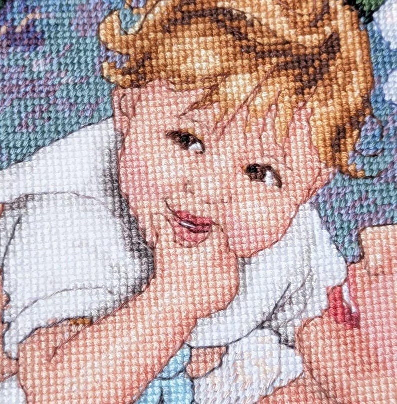 May include: A cross-stitch portrait of a smiling child with light brown hair and rosy cheeks. The child is wearing a white garment and is touching their face. The background is a mix of blue, green, and purple.