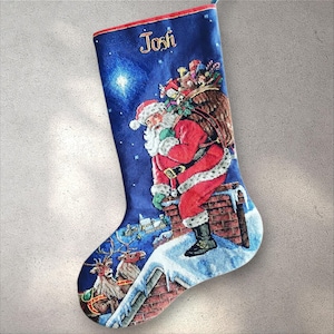 May include: A Christmas stocking with a blue background features Santa Claus on a chimney, with reindeer and a sleigh below. The word "Josh" is embroidered in gold. The stocking is trimmed in red.