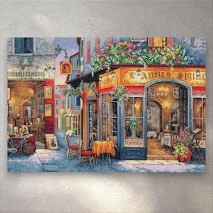 Peut inclure: Une broderie représentant une scène de rue européenne avec un restaurant nommé "L'Amico Sighio" et une librairie. L'œuvre d'art présente une palette de couleurs vives, avec des fleurs, des tables et des chaises. Un vélo est garé près de la librairie.