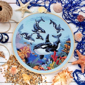 Puede incluir: Un bastidor de bordado de punto de cruz redondo con una escena marina con orcas. La obra de arte representa varias orcas en blanco y negro nadando en un océano azul, rodeadas de coloridos corales y pequeños peces. El bastidor se encuentra sobre un fondo de conchas y elementos náuticos.