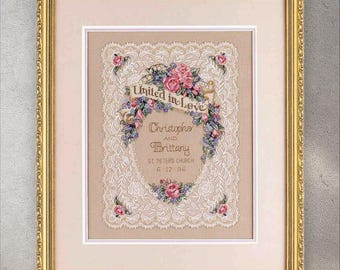 May include: Framed embroidered artwork featuring a cream-colored lace design with floral accents. The artwork includes the text "United in Love" and the names "Christopher and Brittany" with a date. The frame is gold.