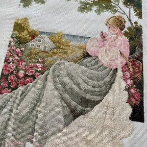May include: A completed needlepoint tapestry depicting a woman in a pink dress seated near a garden. The scene includes a house, flowers, and trees, all rendered in various shades of green, pink, and gray. The tapestry is on a white background.
