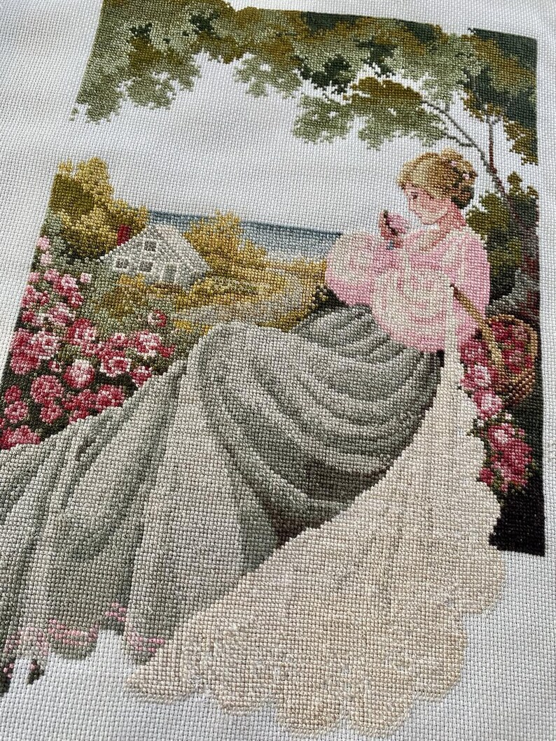 May include: A completed needlepoint tapestry depicting a woman in a pink dress seated near a garden. The scene includes a house, flowers, and trees, all rendered in various shades of green, pink, and gray. The tapestry is on a white background.