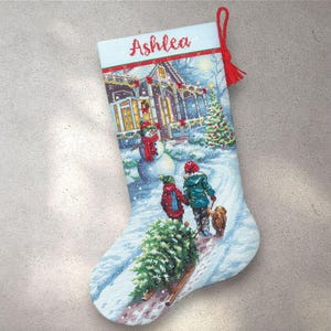Peut inclure: Une chaussette de Noël avec une scène hivernale représentant une maison, un bonhomme de neige, des enfants et un chien. Le nom "Ashlea" est brodé en rouge. La chaussette a un pompon et une boucle rouges pour l'accrocher.