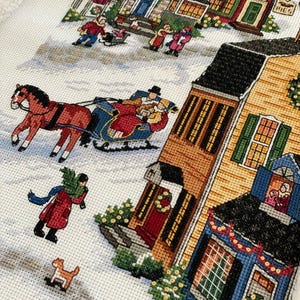 May include: A cross-stitch depicting a winter scene with a horse-drawn sleigh, people, and houses. The houses are yellow, brown, and blue, with Christmas decorations. A person carries a Christmas tree. A small dog runs in the snow.