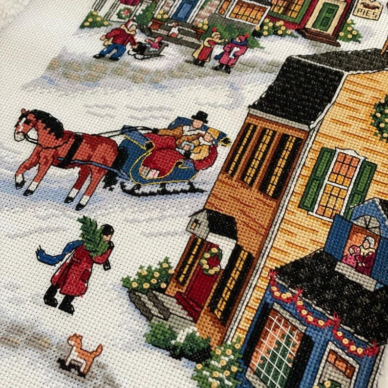 May include: A cross-stitch depicting a winter scene with a horse-drawn sleigh, people, and houses. The houses are yellow, brown, and blue, with Christmas decorations. A person carries a Christmas tree. A small dog runs in the snow.