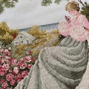 May include: Needlepoint tapestry depicting a woman in a pink dress seated near a garden. The scene includes a house, trees, and a body of water. The tapestry features a variety of colors, including pink, green, and gray, and is likely a vintage piece.