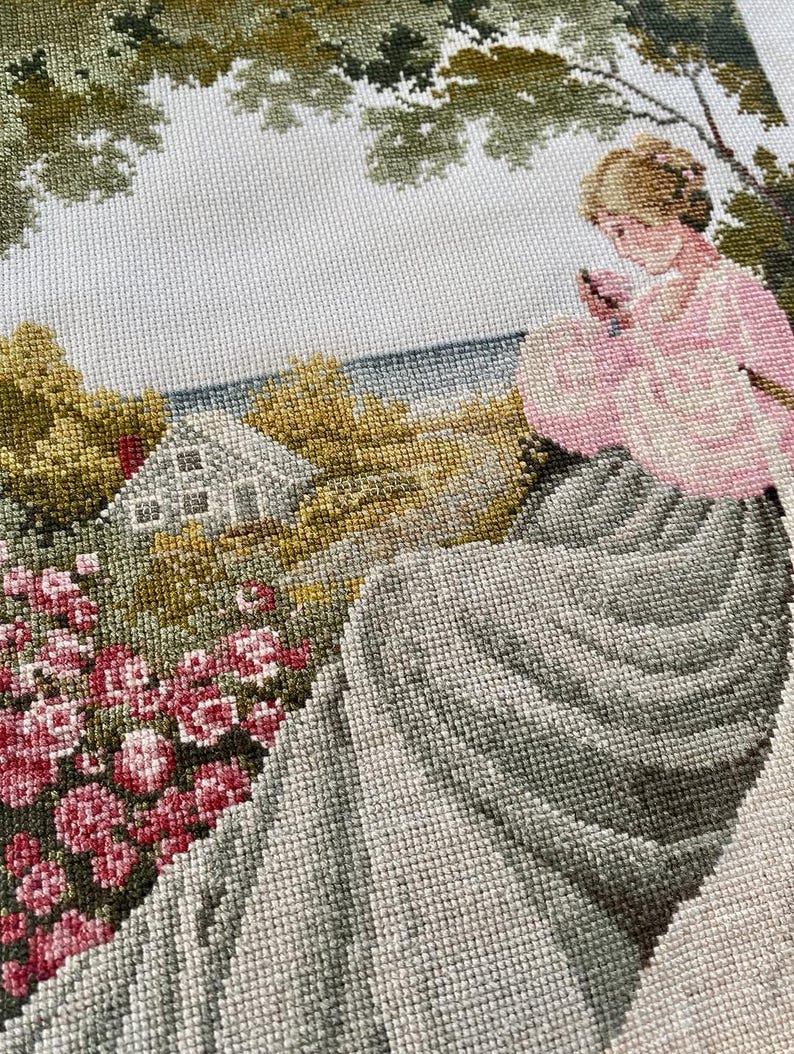 May include: Needlepoint tapestry depicting a woman in a pink dress seated near a garden. The scene includes a house, trees, and a body of water. The tapestry features a variety of colors, including pink, green, and gray, and is likely a vintage piece.