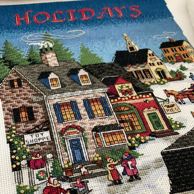 May include: A cross-stitch textile featuring a winter holiday scene. The word "HOLIDAYS" is stitched across the top. The scene depicts snow-covered houses, people, and a horse-drawn carriage. One building is labeled "TOY SHOPPE".