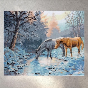 May include: A cross-stitch artwork depicting two horses in a winter landscape. One horse is gray, drinking from a stream, while the other is brown and stands nearby. The scene includes snow-covered banks, trees, and a soft, diffused light.